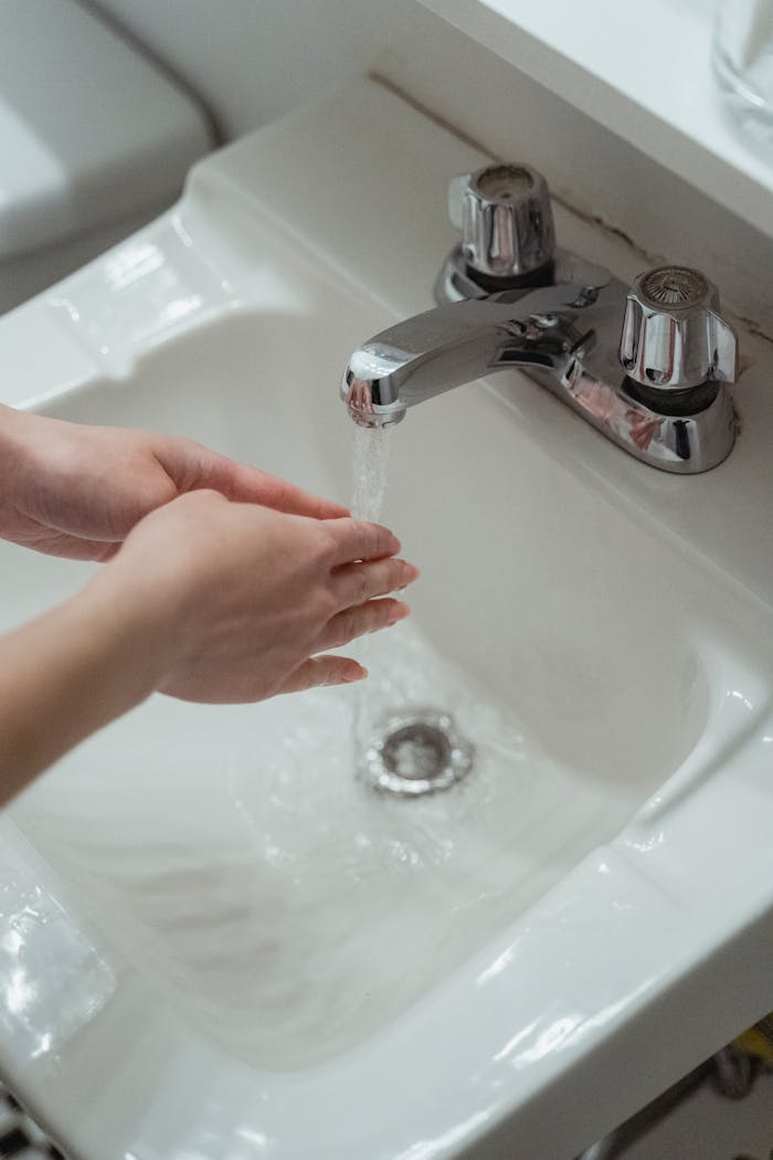 services-01 Close-up of hands being washed under a modern stainless steel faucet in a white washbasin.
